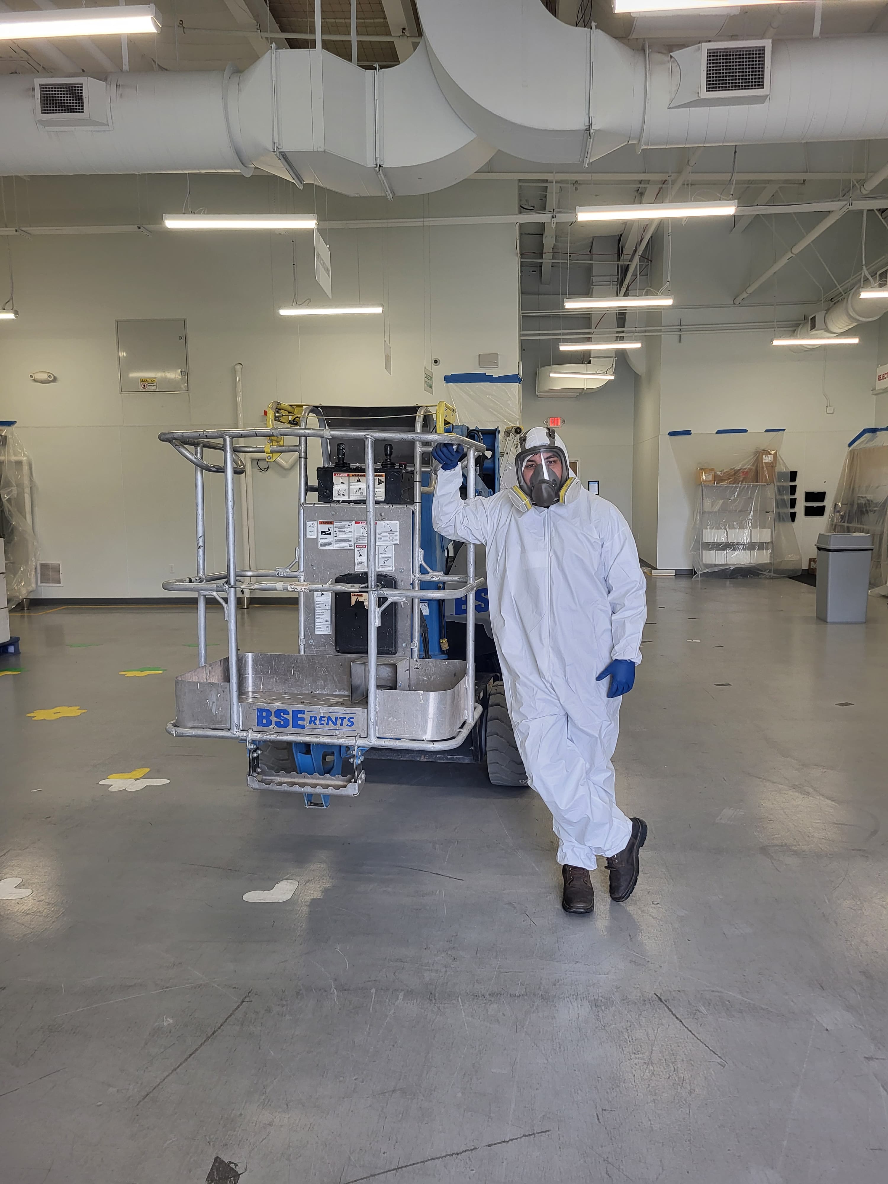 Worker in white hazmat suit and respirator standing next to industrial lift equipment.