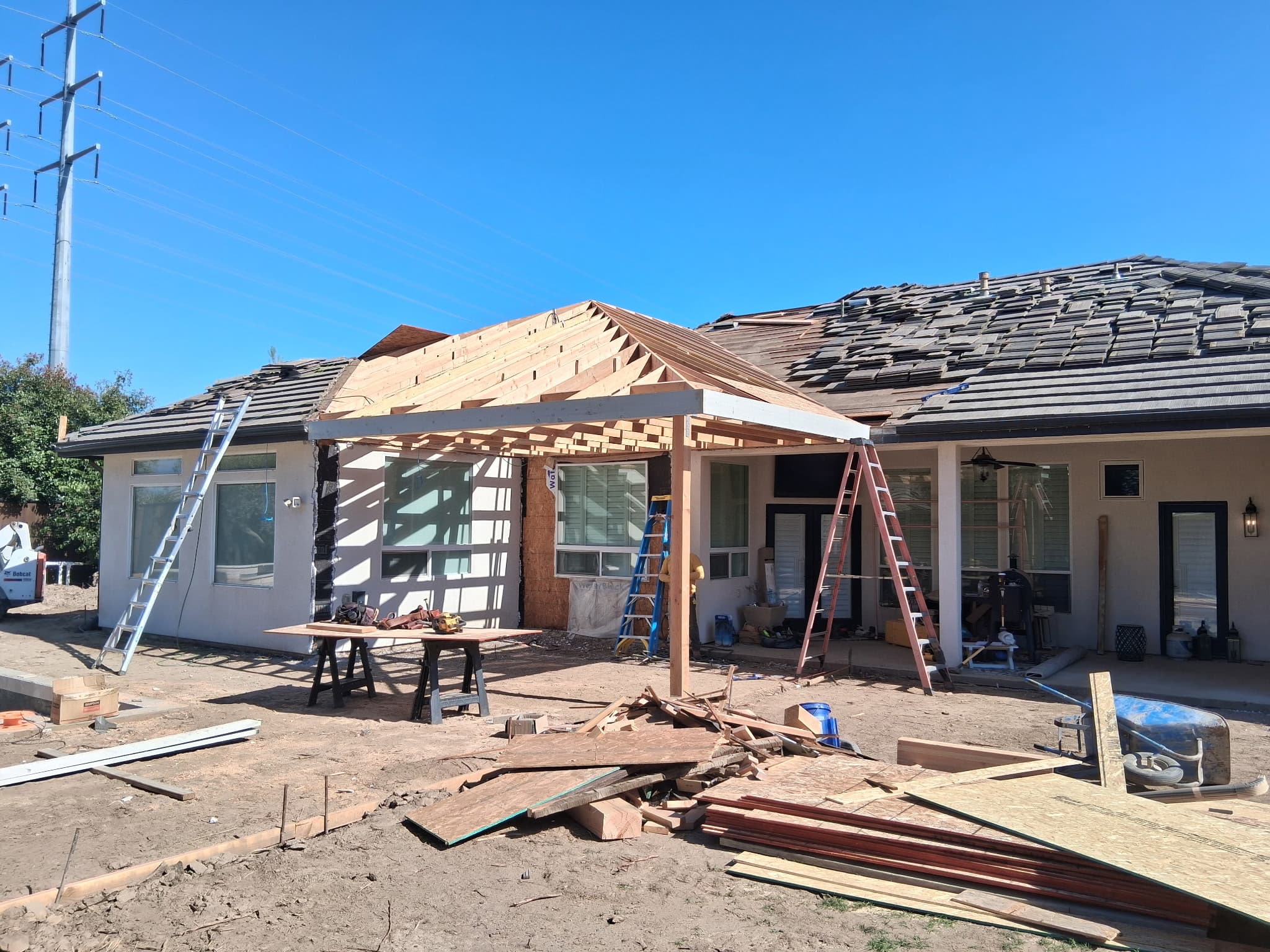 House under renovation featuring a new wooden patio roof frame, ladders, and construction debris.