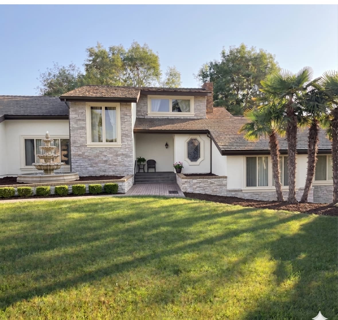 Stone and stucco house with a tiered fountain, palm trees, and a sunlit green lawn.