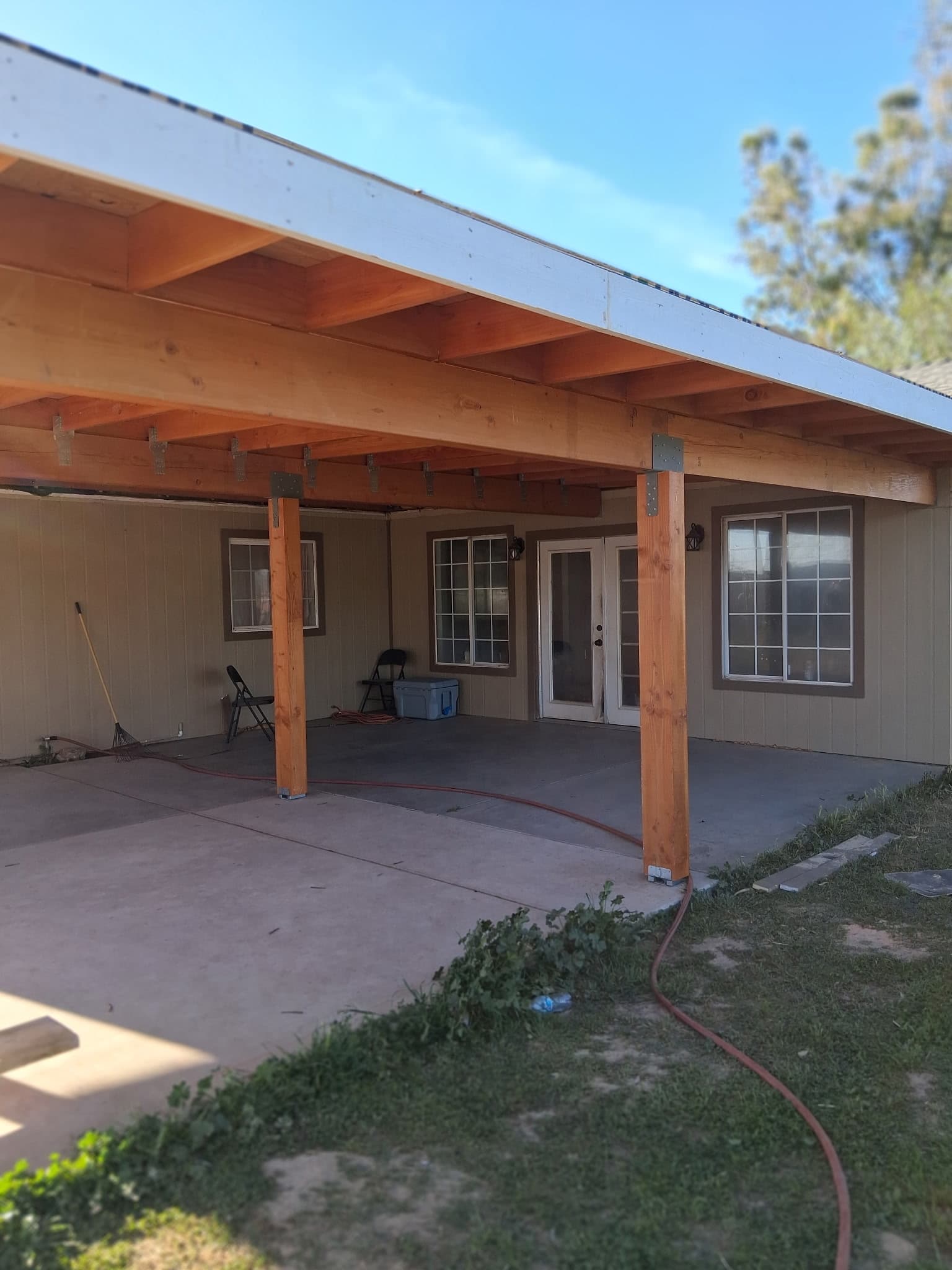 Wooden patio cover with thick beams and posts over a concrete patio by a house.