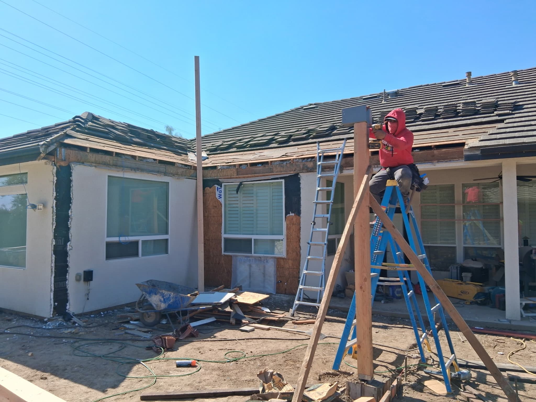 Construction worker in a red hoodie drills a wooden post from a blue ladder.