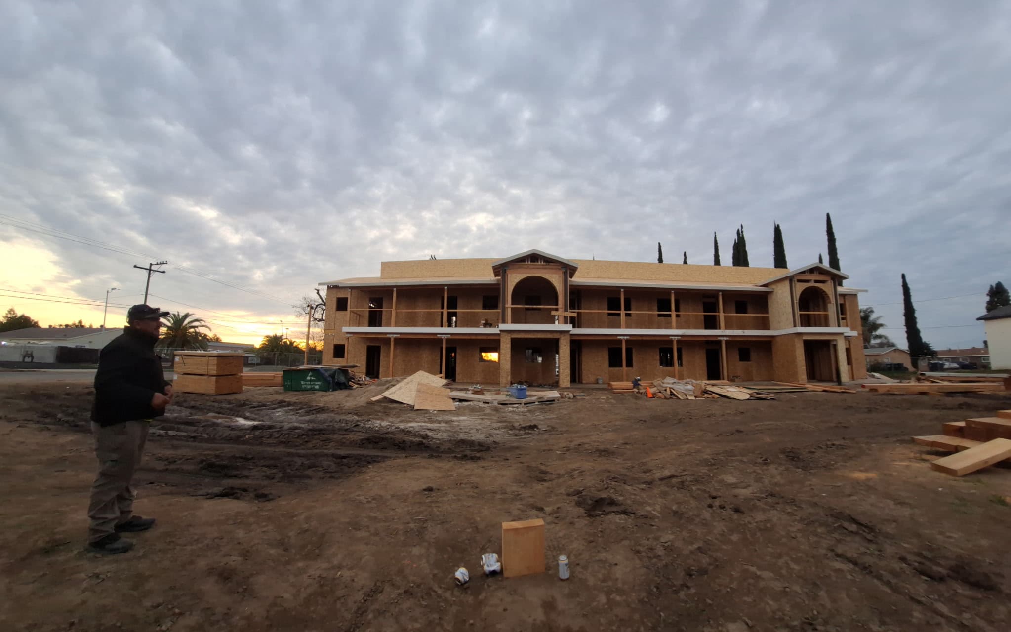 Two-story plywood building under construction on a muddy site under a dramatic cloudy sunset.