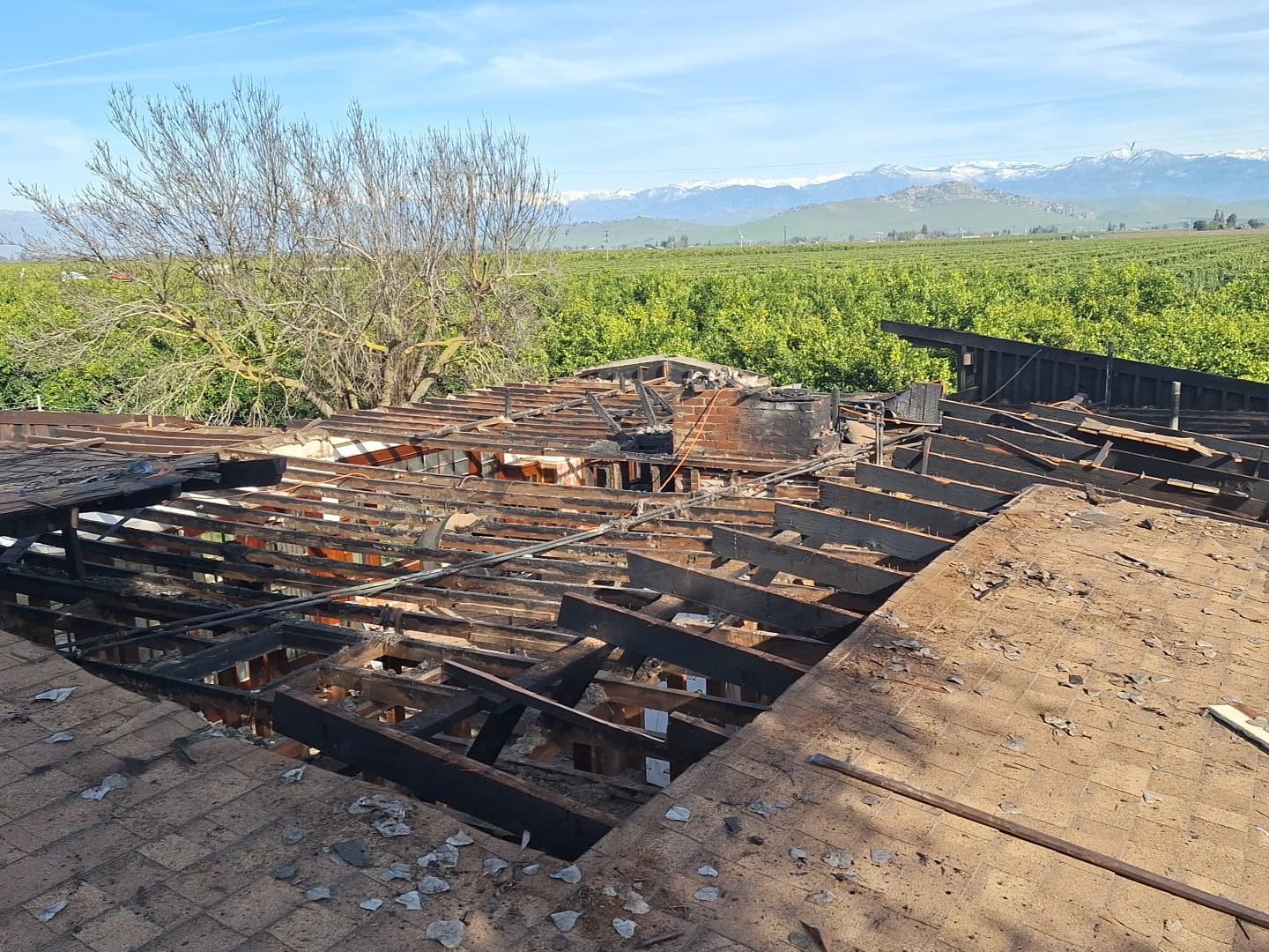 Fire-damaged roof with charred wooden beams overlooking green orchards and distant snow-capped mountains.